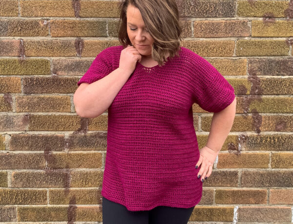 A woman stands in front of a brick wall, wearing a hand crocheted vneck tunic shirt.