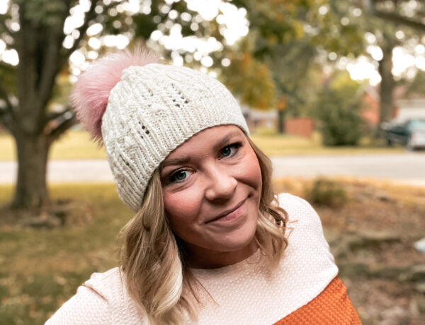 A woman stands outside, wearing a handmade knit beanie with faux fur pom pom