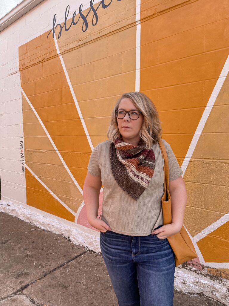 A woman stands in from of a mural, wearing a tunisian crochet cowl in various colors.